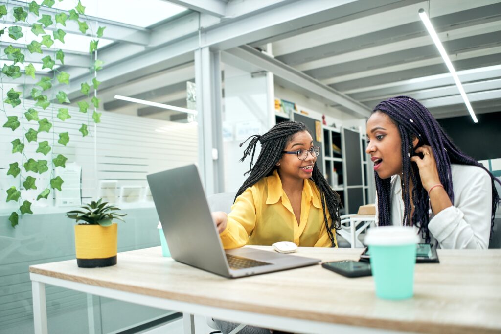 Two young women in office looking surprised at something they are seeing on laptop screen.